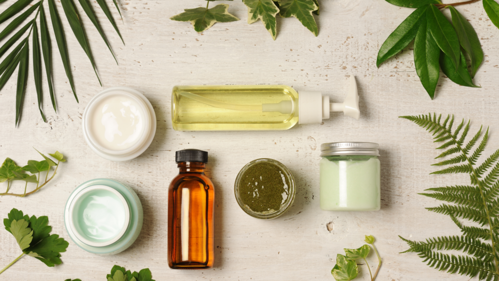 Vue aérienne d’un assortiment de produits de soins naturels sur une table en bois clair, entourés de feuilles vertes. Les produits incluent des pots de crèmes, une bouteille d’huile, un flacon ambré, et un petit pot de masque vert, évoquant un univers naturel et apaisant.