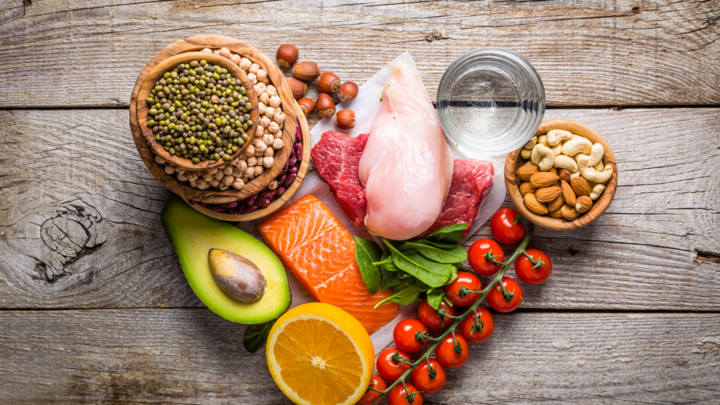 Une sélection variée d'aliments sains disposés sur une surface en bois : des légumes comme l'avocat, des tomates cerises, des protéines comme du poulet, du bœuf et du saumon, des légumineuses, des fruits secs (amandes, noix de cajou) et une orange, accompagnés d'un verre d'eau.
