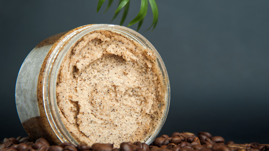 Un pot ouvert contenant un exfoliant naturel granuleux de couleur beige clair, posé sur une surface décorée de grains de café bruns. Une feuille verte est visible en arrière-plan, donnant une ambiance naturelle et apaisante.