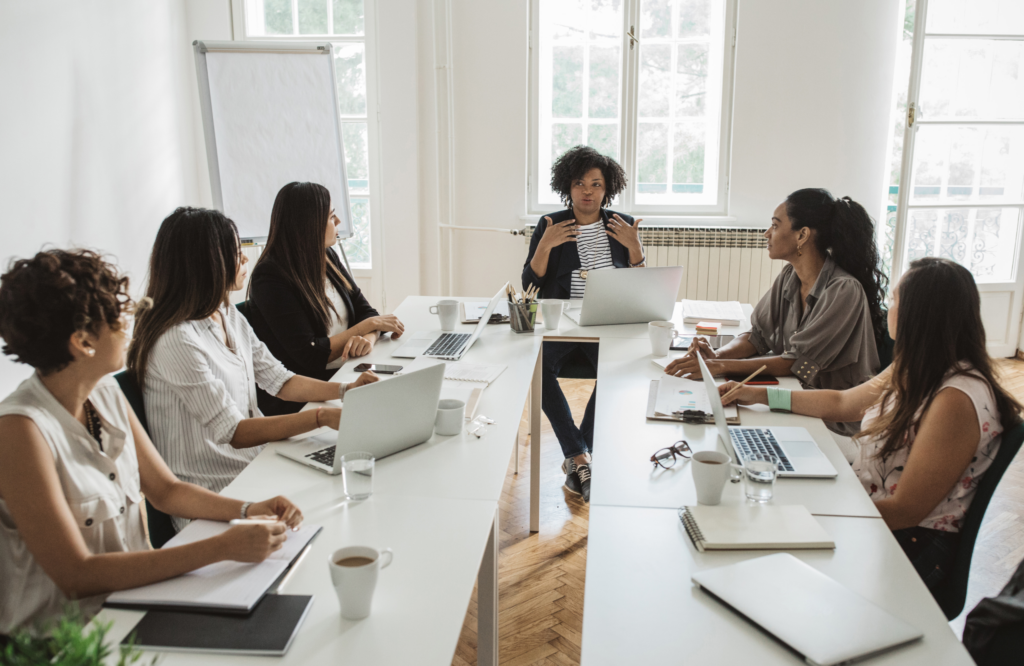 Six femmes sont assises autour d'une table blanche dans une salle lumineuse. Elles discutent, certaines prennent des notes et d'autres utilisent des ordinateurs portables. Une femme au centre, en train de parler, semble diriger la réunion.