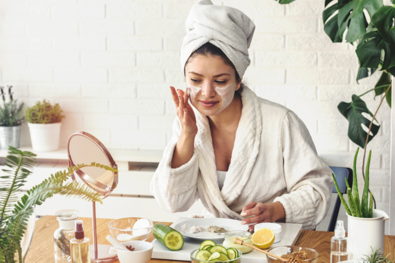 L’image montre une femme assise à une table, portant un peignoir blanc et une serviette enroulée autour de ses cheveux. Elle applique une crème sur son visage tout en regardant un miroir posé devant elle. Sur la table, on voit des ingrédients naturels comme des tranches de concombre, un demi-citron, et plusieurs bols contenant des produits faits maison. En arrière-plan, des plantes décorent la pièce, créant une ambiance fraîche et naturelle.