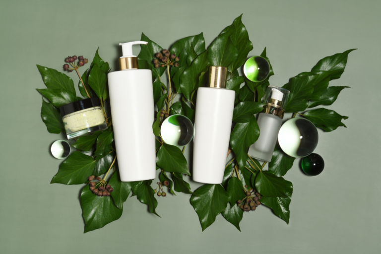 L'image montre des produits de soins de la peau posés sur des feuilles vertes : deux grands flacons blancs, un pot de crème et une petite bouteille avec un distributeur. Des sphères en verre décorent la scène, créant une ambiance naturelle et élégante.