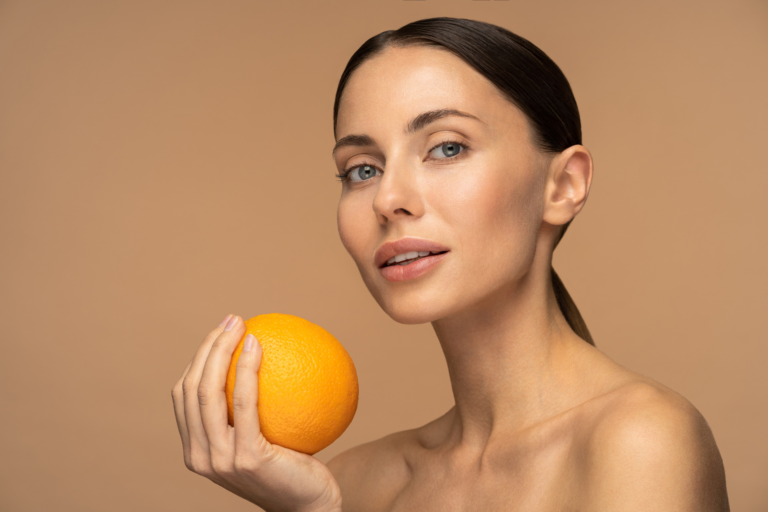 L’image montre une femme à la peau claire qui tient une orange dans sa main. Elle a les cheveux tirés en arrière et un léger sourire. Le fond beige uni donne une ambiance naturelle et apaisante. L’orange illustre le lien entre alimentation et santé de la peau.