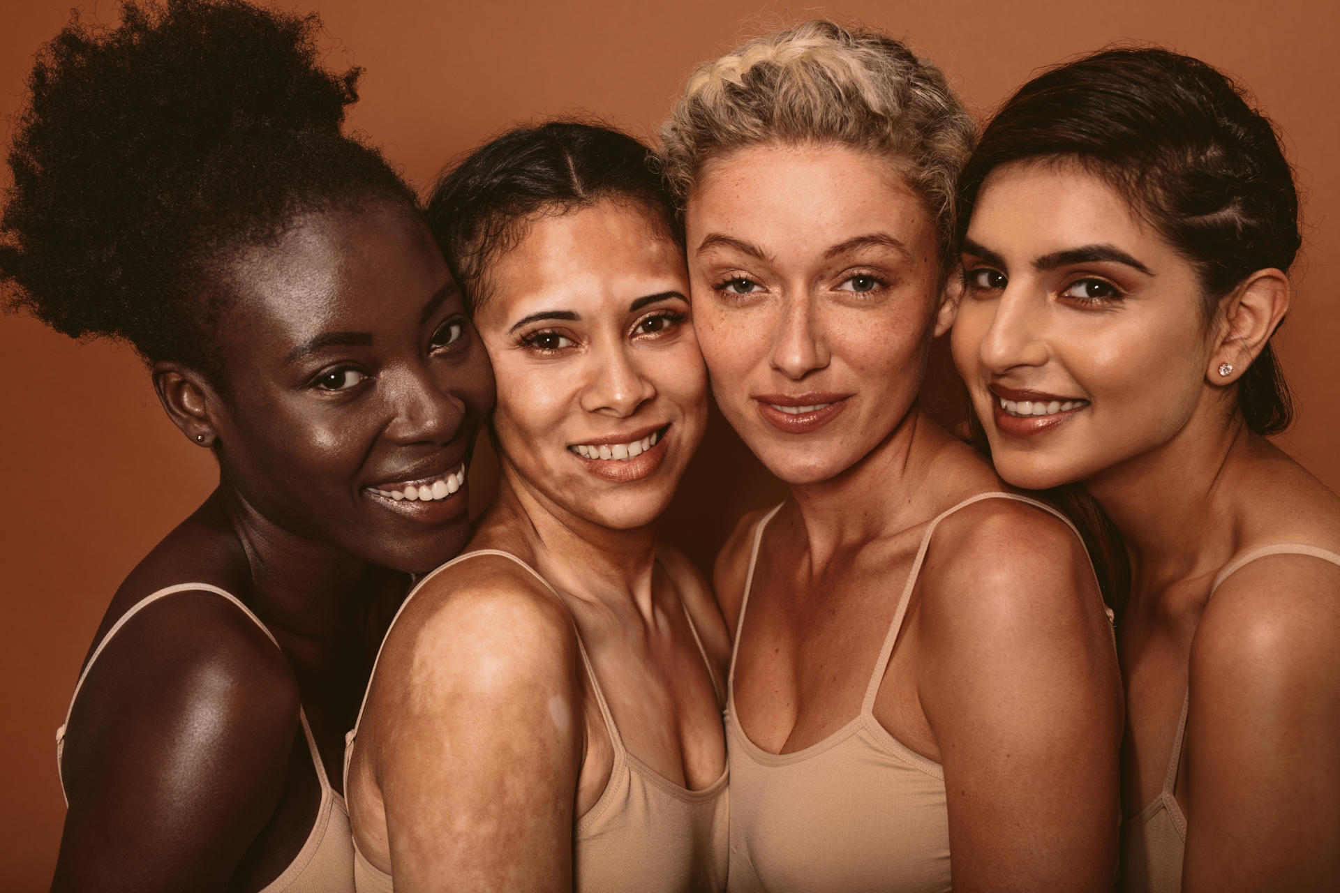 L’image montre quatre femmes souriantes, proches les unes des autres. Elles ont des peaux et des traits différents, reflétant la diversité. Elles portent des hauts beige clair, et le fond est marron chaud. L’ensemble dégage une impression de beauté naturelle et d’harmonie.