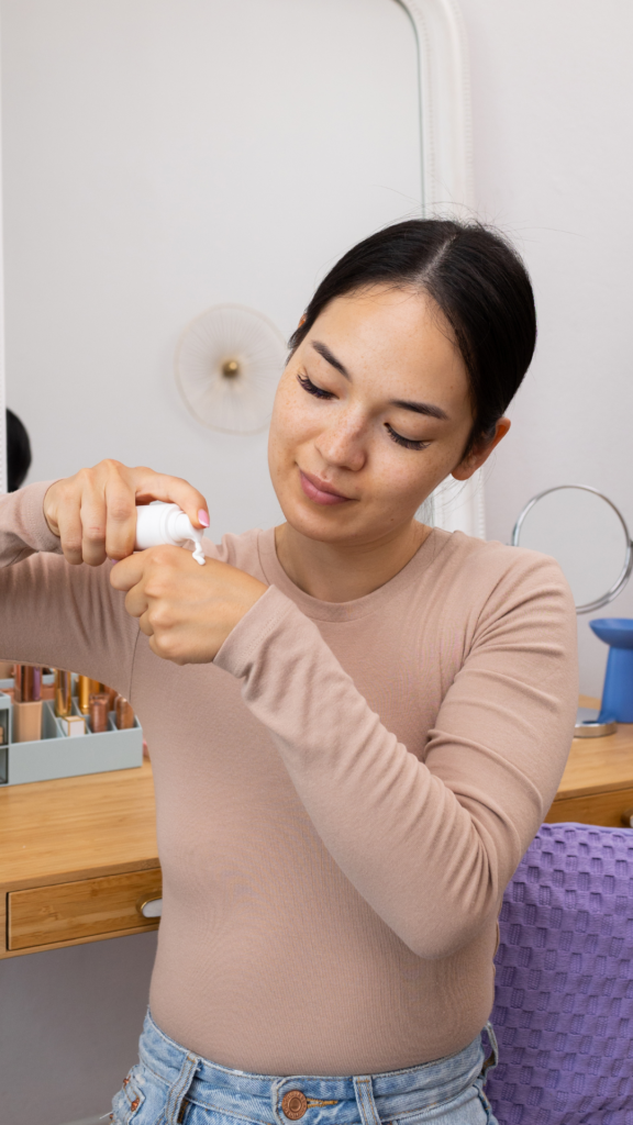 Une femme portant un haut beige et un jean applique une crème sur le dos de sa main. Elle est assise devant une coiffeuse en bois, avec des produits de beauté rangés dans un organisateur et un miroir en arrière-plan.