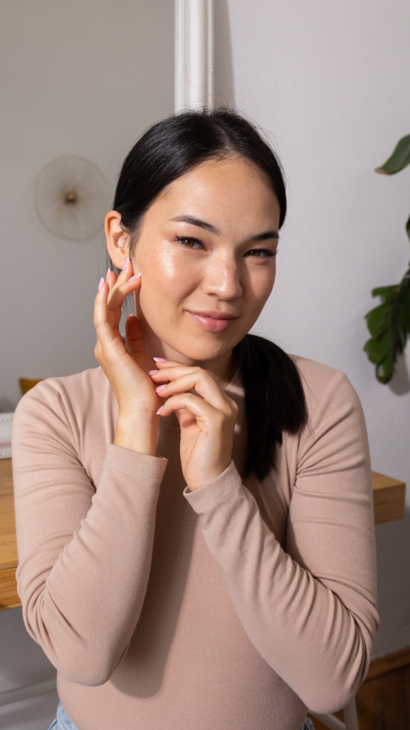 Une femme souriante portant un haut beige et un jean touche délicatement sa joue avec ses mains. Elle est assise devant une coiffeuse en bois, avec une plante et un miroir visibles en arrière-plan.
